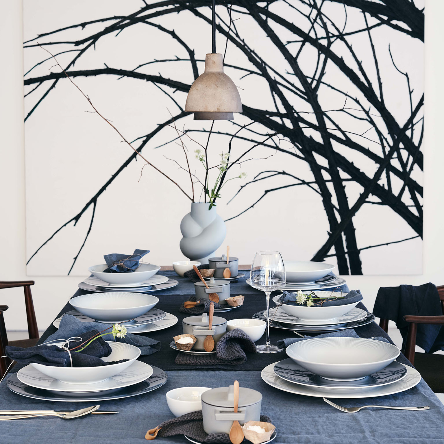 Vue latérale d'une table entièrement dressée pour six personnes avec des assiettes à soupe et des assiettes plates TAC Sensual, des serviettes en lin, des accessoires de table, un vase Node et un grand tableau en arrière-plan.